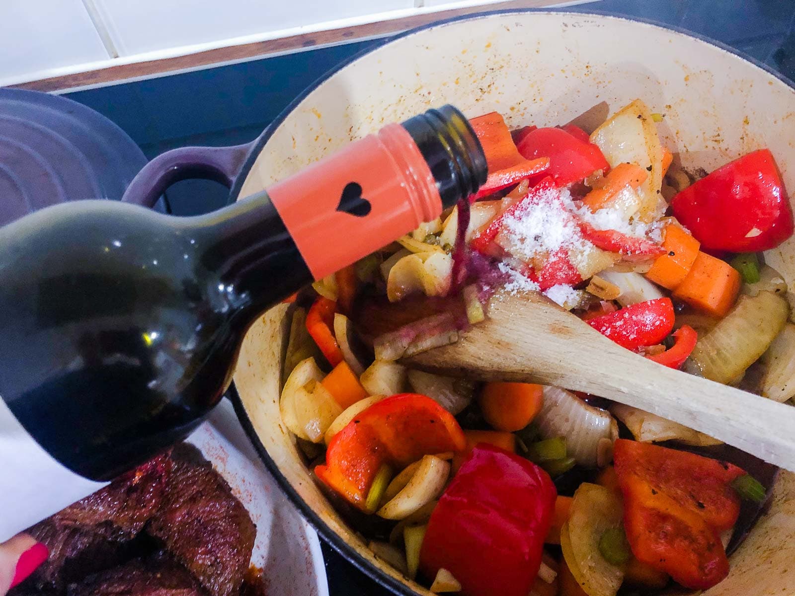 A hand adding red wine from a bottle to chopped vegetables in preparation of making a paprika beef casserole with the diced beef just at the bottom ready to add to the casserole.