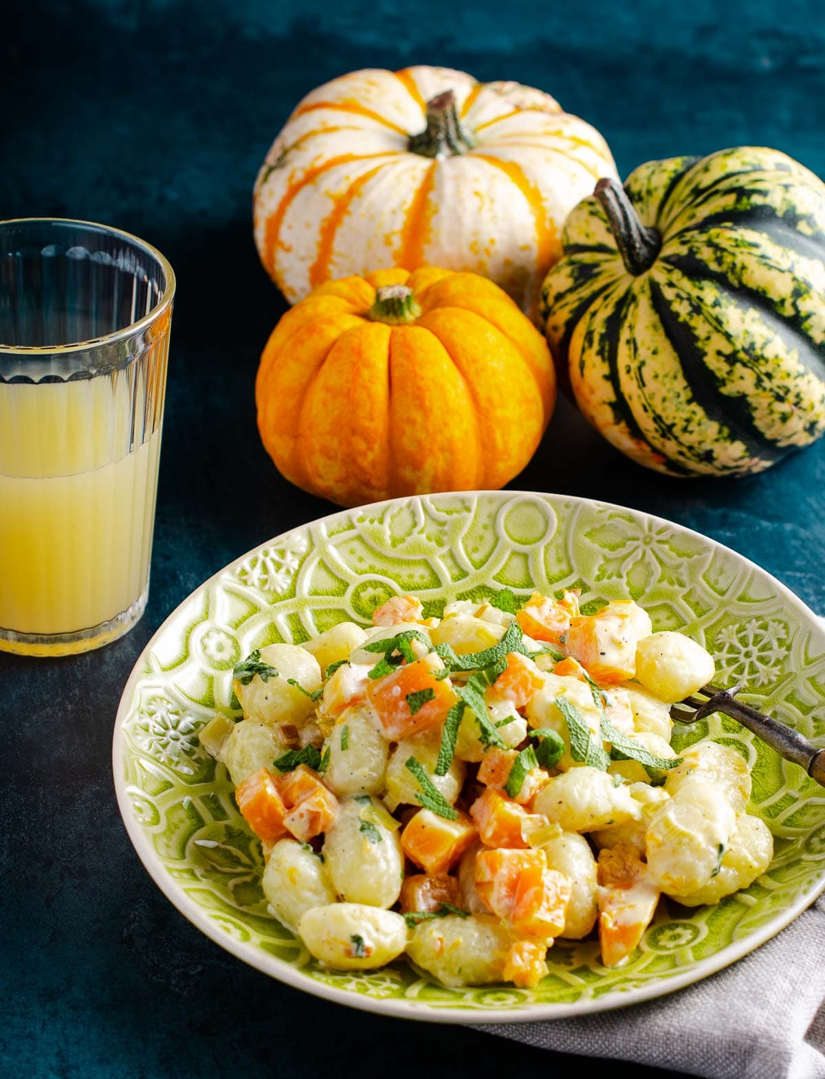 A plate of creamy gnocchi with pan fried pumpkin pieces and sage served an an ornate green plate, with a glass of orange juice in the back and a mix of orange and green pumpkins to the back.