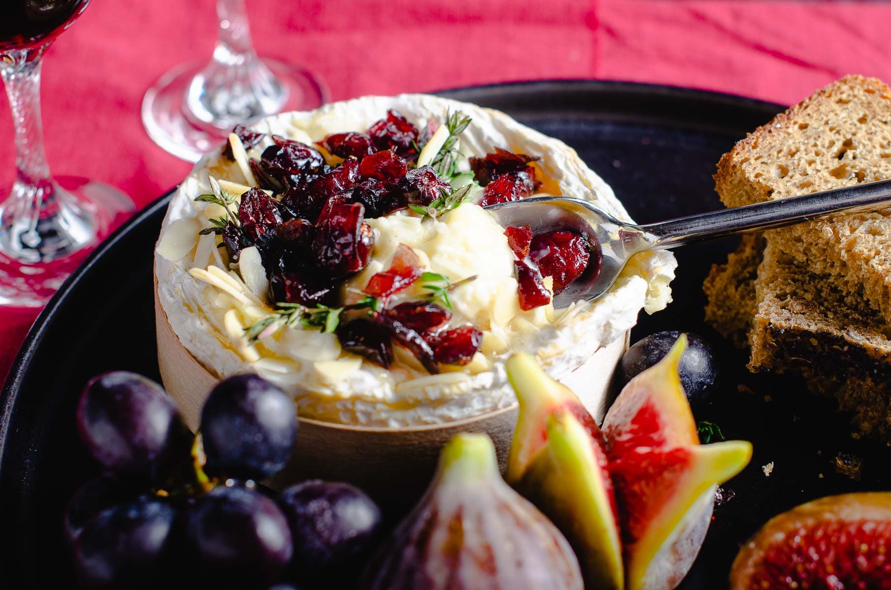 A decadent cheeseboard served with a whole baked cheese topped with cranberries, thyme and almonds and served with fresh figs, black grapes and a nutty brown bread.
