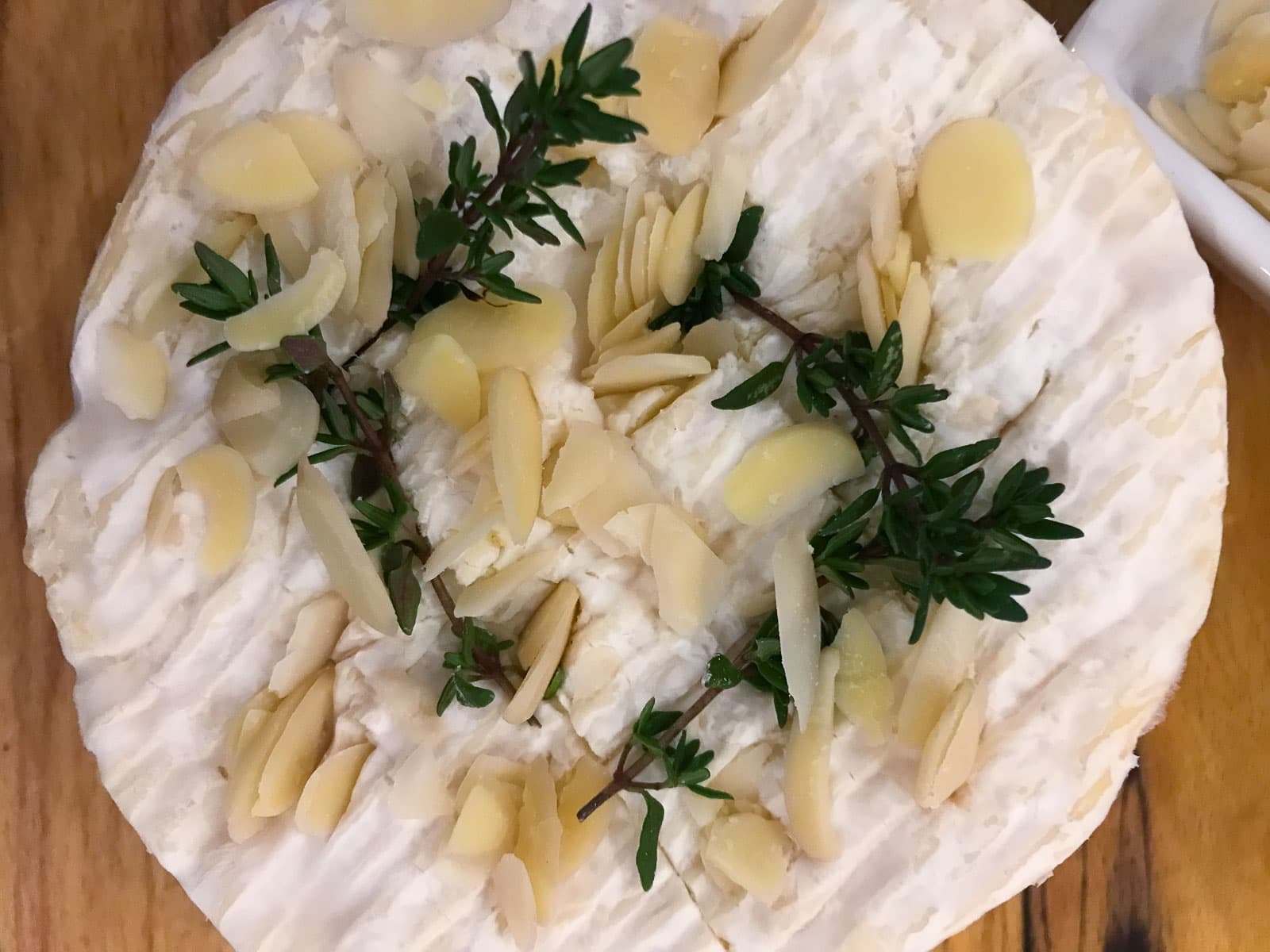 A whole camembert stuffed with fresh thyme leaves, sliced almonds on a wooden board.