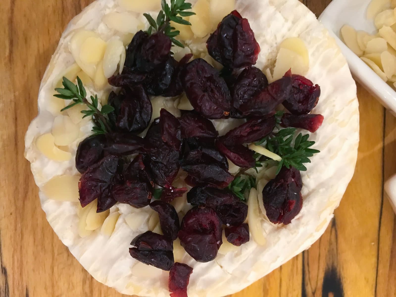 A whole Camembert cheese with toppings of dried cranberries, sliced almonds and thyme to be baked.