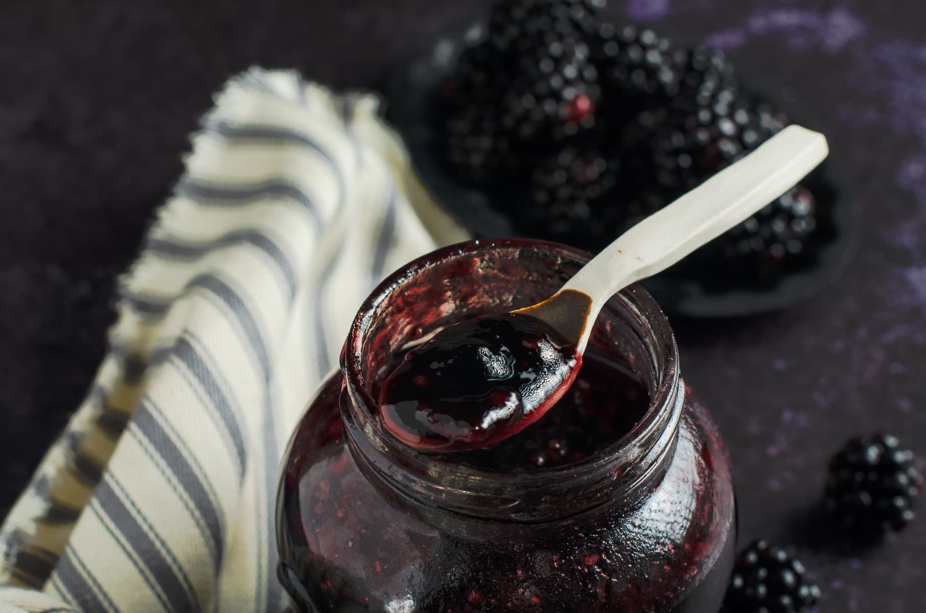 A round jar of blackberry jelly with a small ceramic spoon on the top, some fresh blackberries to the back and side and a striped French linen on a dark background.