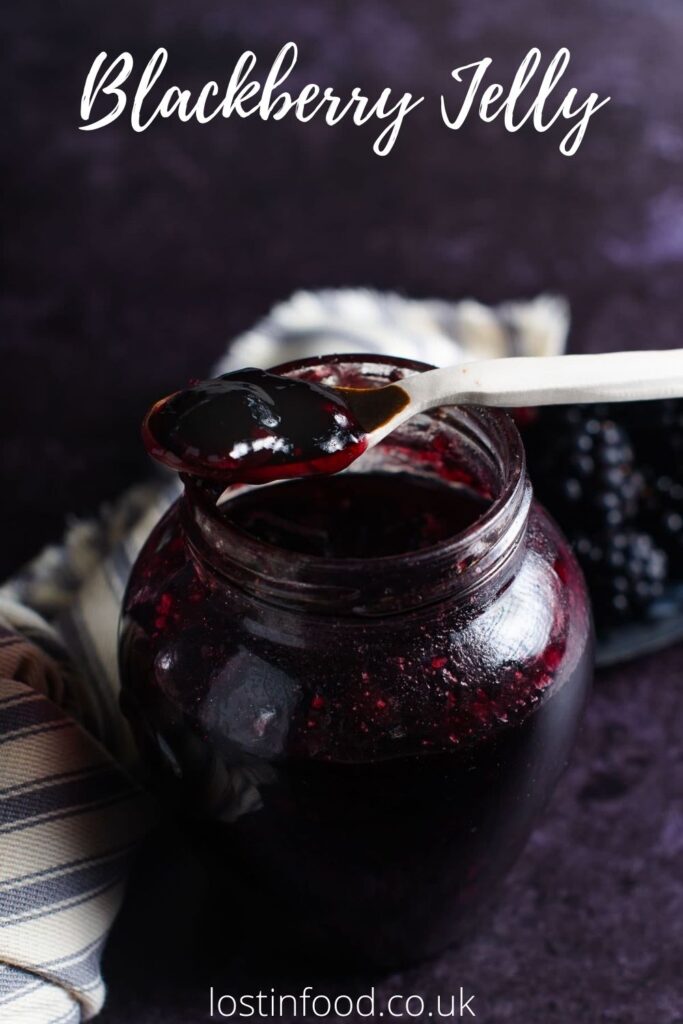 A Pinterest graphic of a round jar blackberry jelly with a small ceramic spoon on the top, some fresh blackberries to the back and side and a striped French linen on a dark background.
