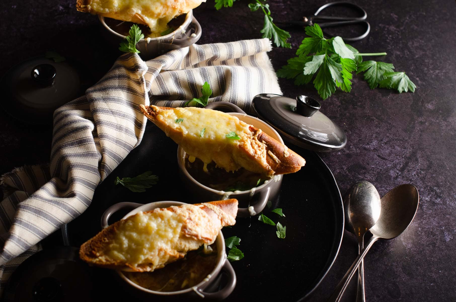 A dark scene of french onion soup bowls topped with toasted bread and melted cheese on a black surface.