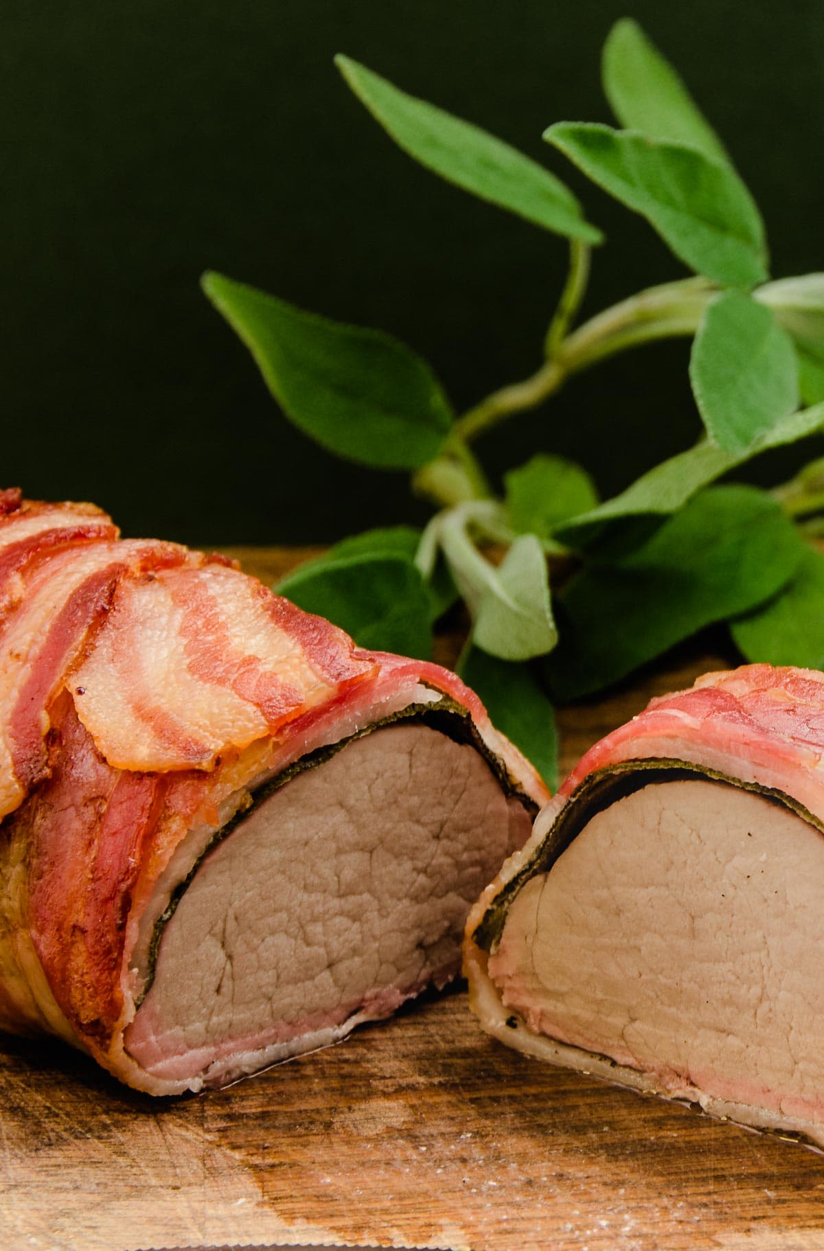 Pork fillet wrapped in bacon and sage sliced in half to show the inside and some fresh sage leaves to the back.