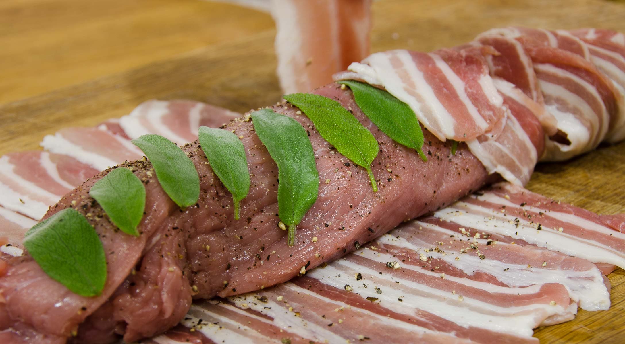 Bacon strips being wrapped around a pork fillet with sage leaves.