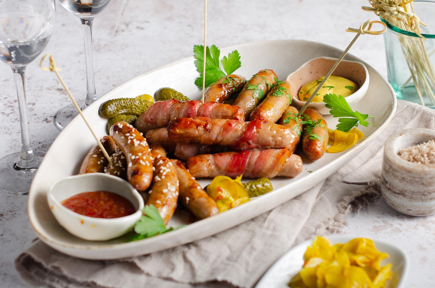 A serving platter of cocktail sausages with 3 types of flavours, dipping sauces, some pickles and cocktail sticks and sparkling wine.