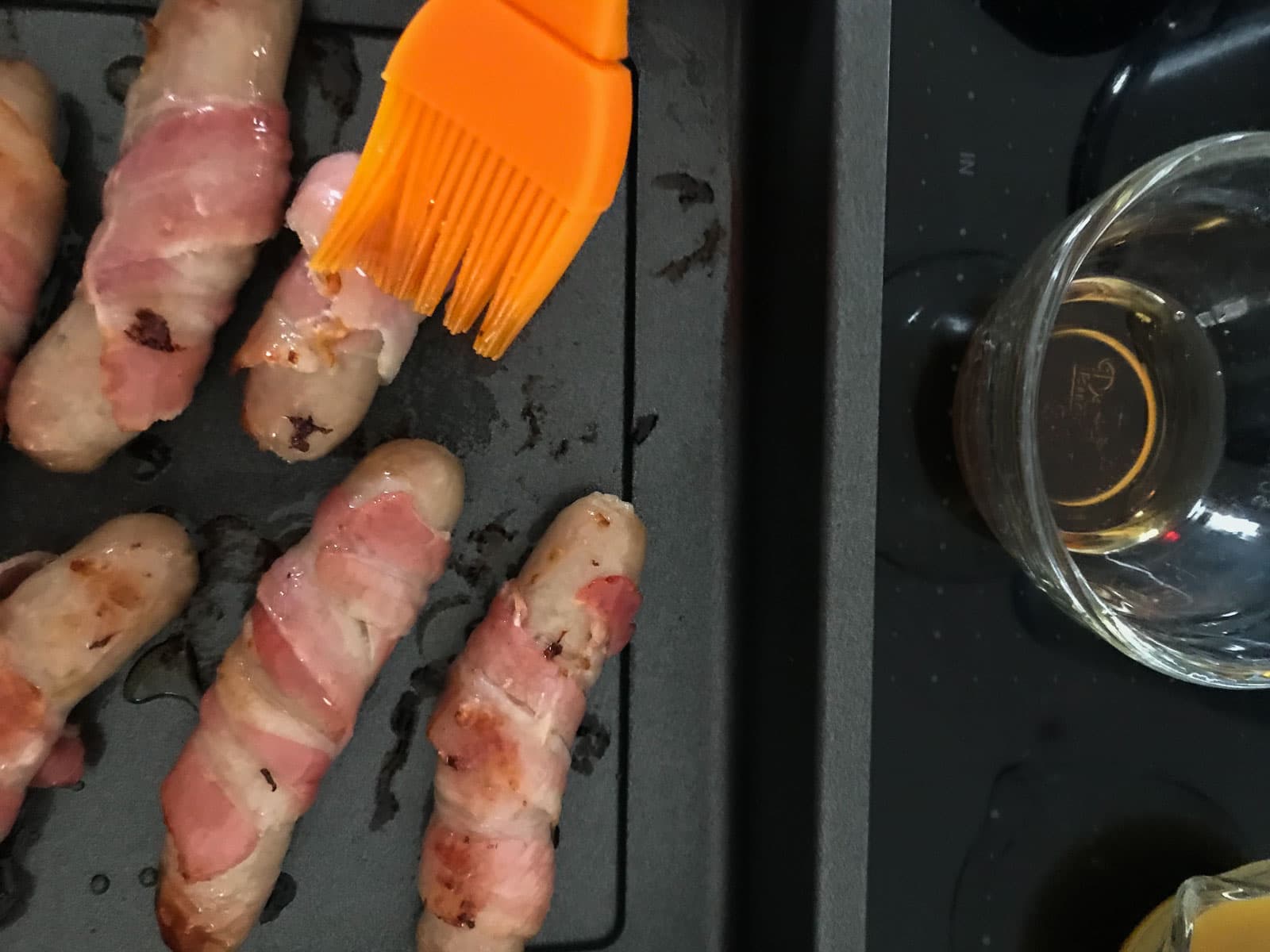 A silicone pastry brush adding maple syrup over part cooked bacon wrapped sausages.
