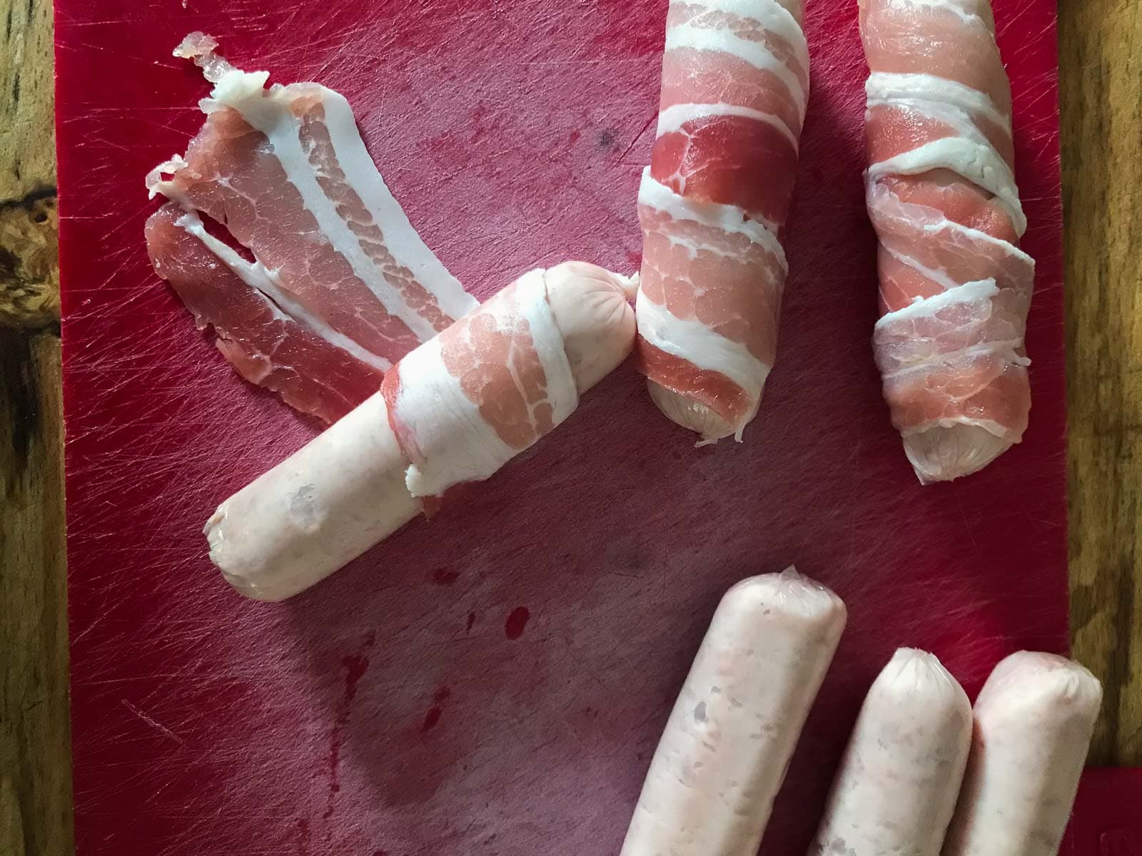 Wrapping sausages in bacon on a red chopping board.