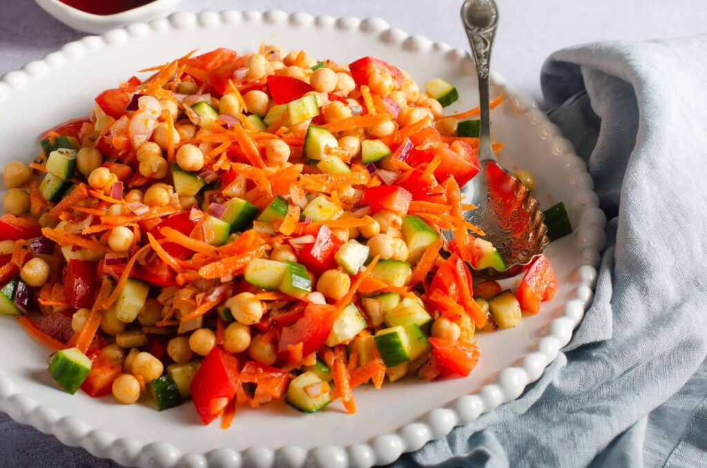 A large plate with chickpea and vegetable dressed salad with a large serving spoon on the side.