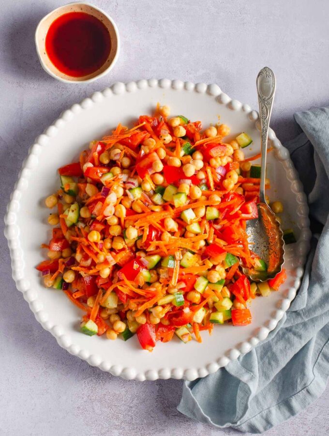 A vibrant plate of chickpea salad with grated carrot, chopped cucumber and tomato on a grey backdrop with a small bowl of harissa dressing to the back.