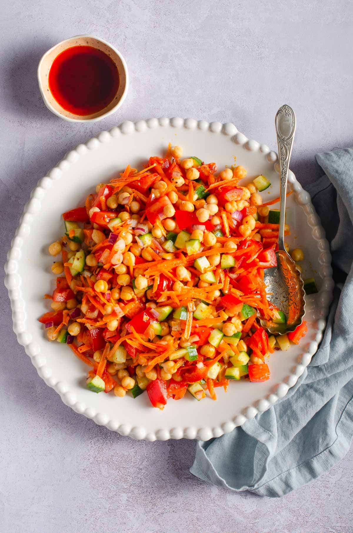 A vibrant plate of chickpea salad with grated carrot, chopped cucumber and tomato on a grey backdrop with a small bowl of harissa dressing to the back.