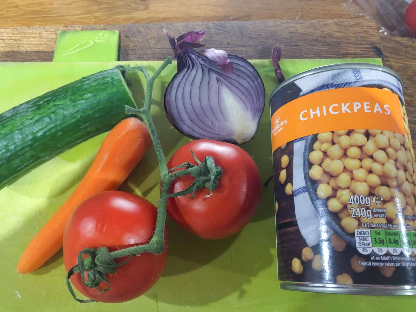 Ingredients on a green chopping board consisting of tomatoes, carrots, cucumber, red onion and a tin of chickpeas.