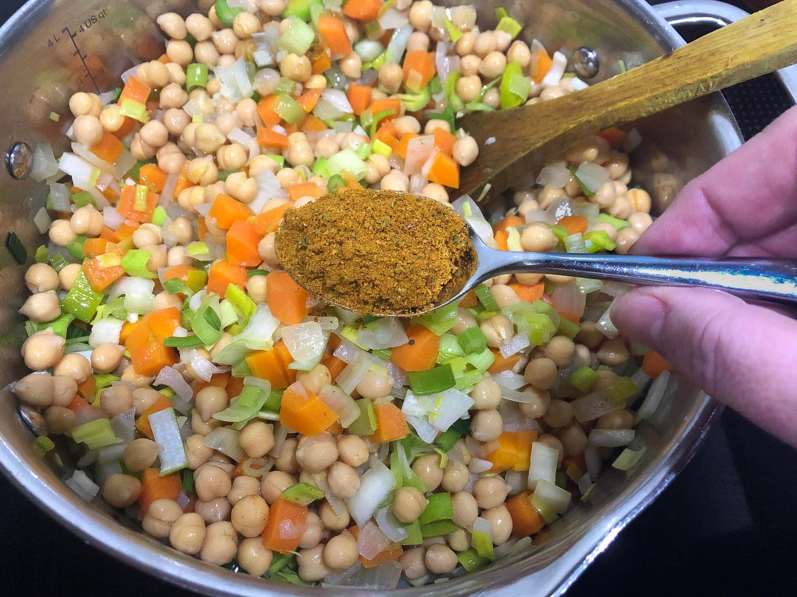 A hand adding a spoon of dried curry powder to sauteed vegetables and chickpeas.