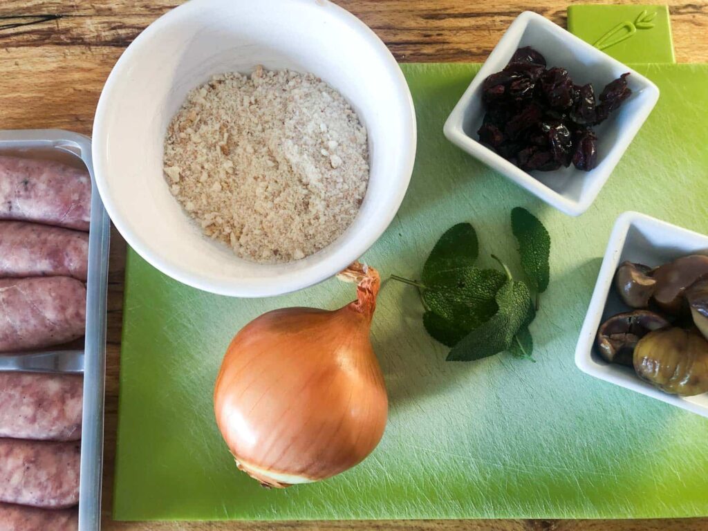 Ingredients laid out to make sausage balls, including an onion, dried breadcrumbs, fresh sage leaves, whole roasted chestnuts and dried cranberries.