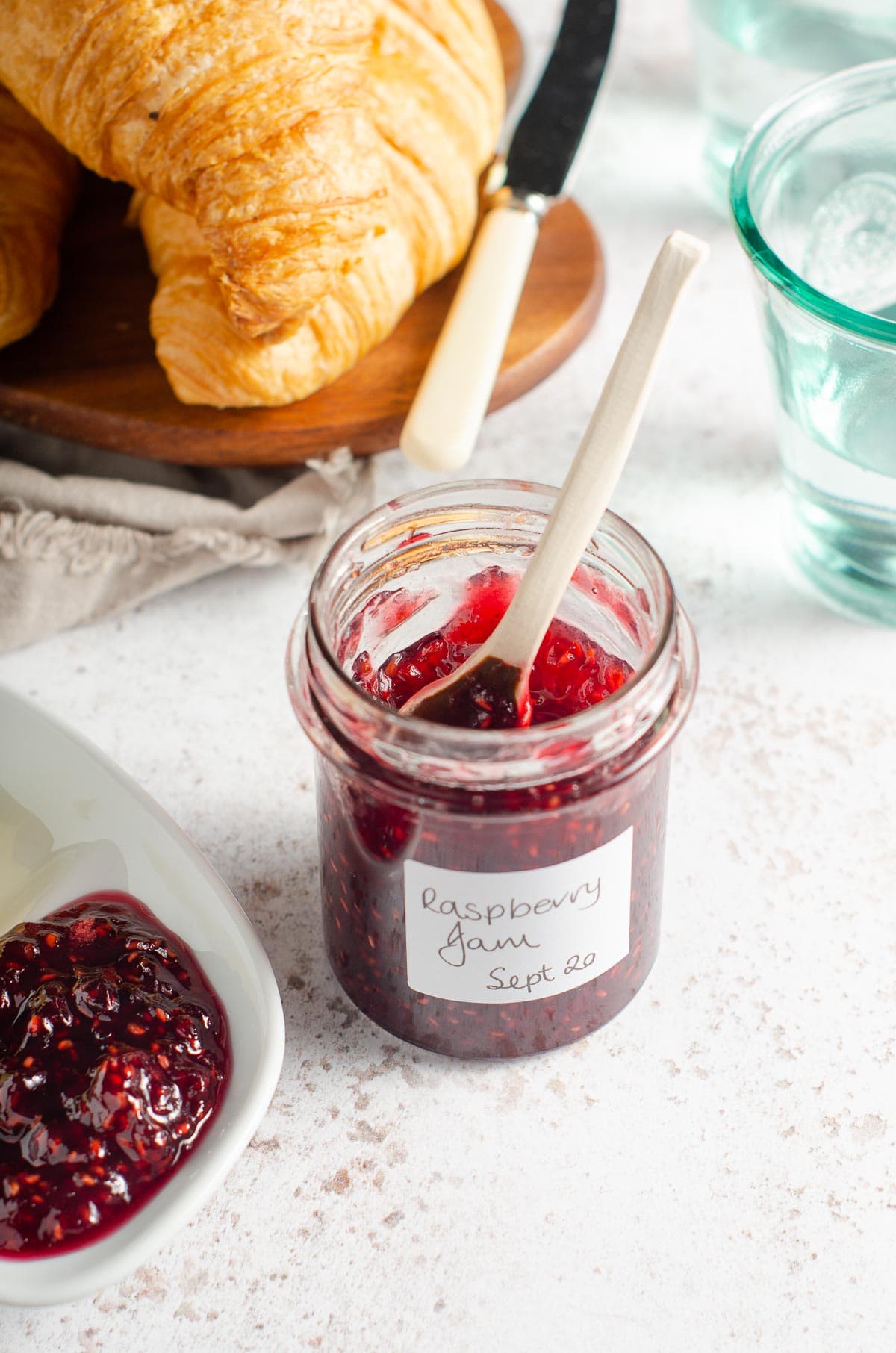 An open jar of homemade raspberry jam served with fresh croissants and butter.
