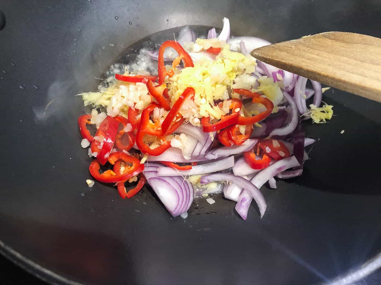red onions, garlic, red chilies and grated ginger being fried in a wok.