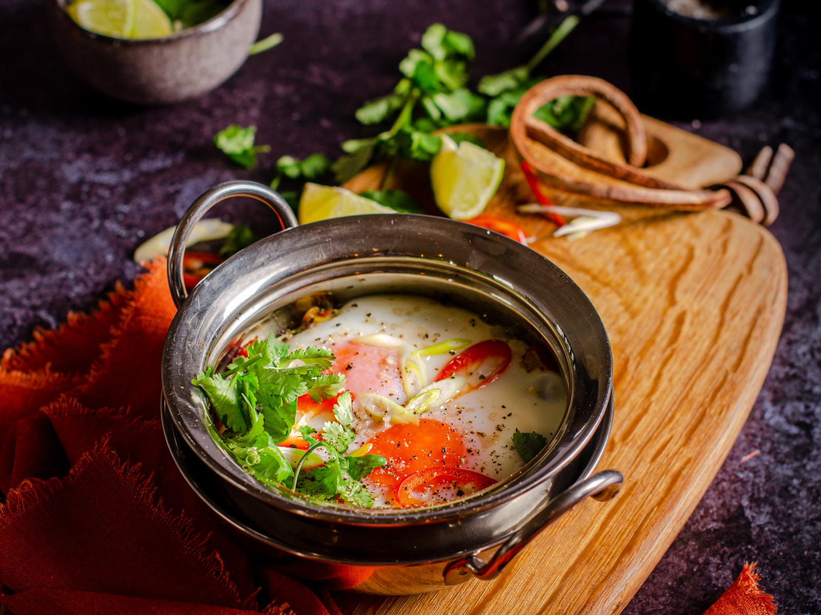 A dish of curried baked eggs, topped with fresh corriander, fresh slices of green onions and chillies served on a wooden board with an orange linen napkin.