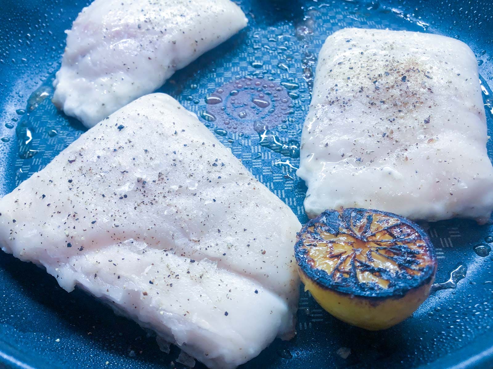 Seasoned hake fillets frying skin side down and lemon halves seared n the pan.