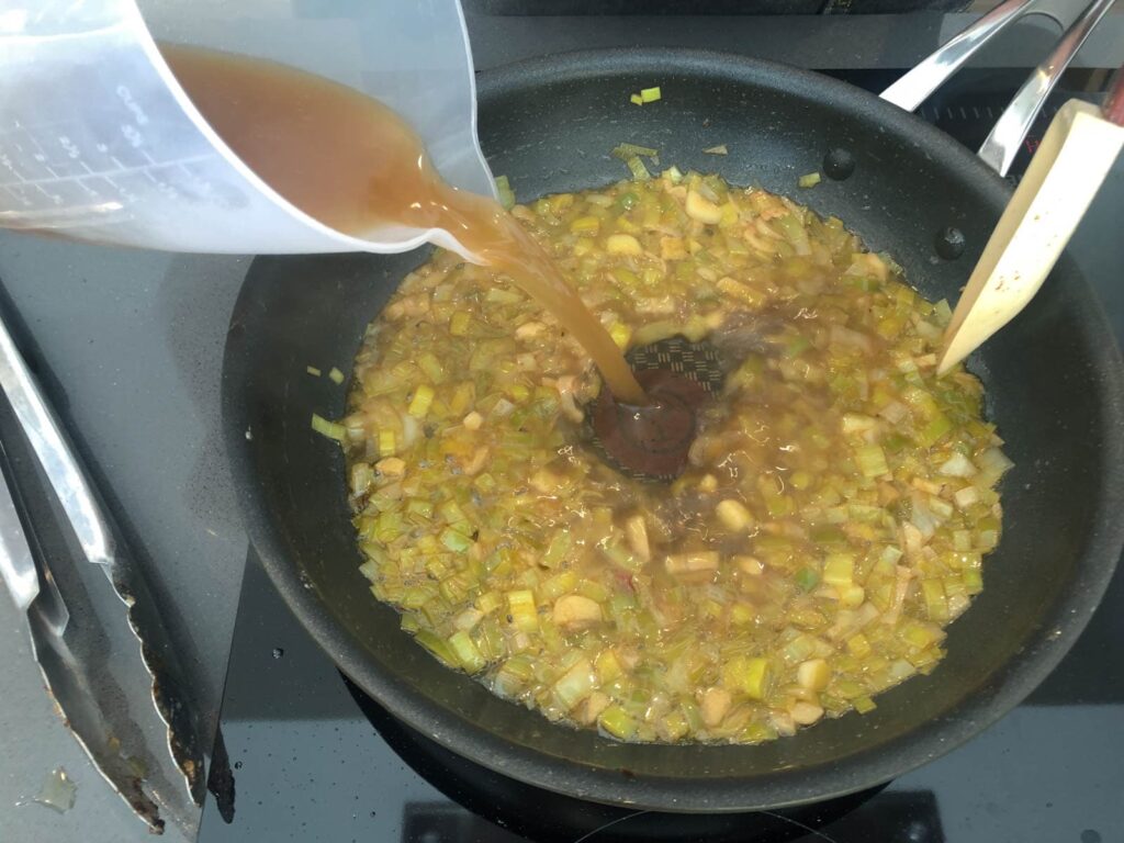 A jug of chicken stock being added to cooked leeks and garlic to make a tarragon sauce.