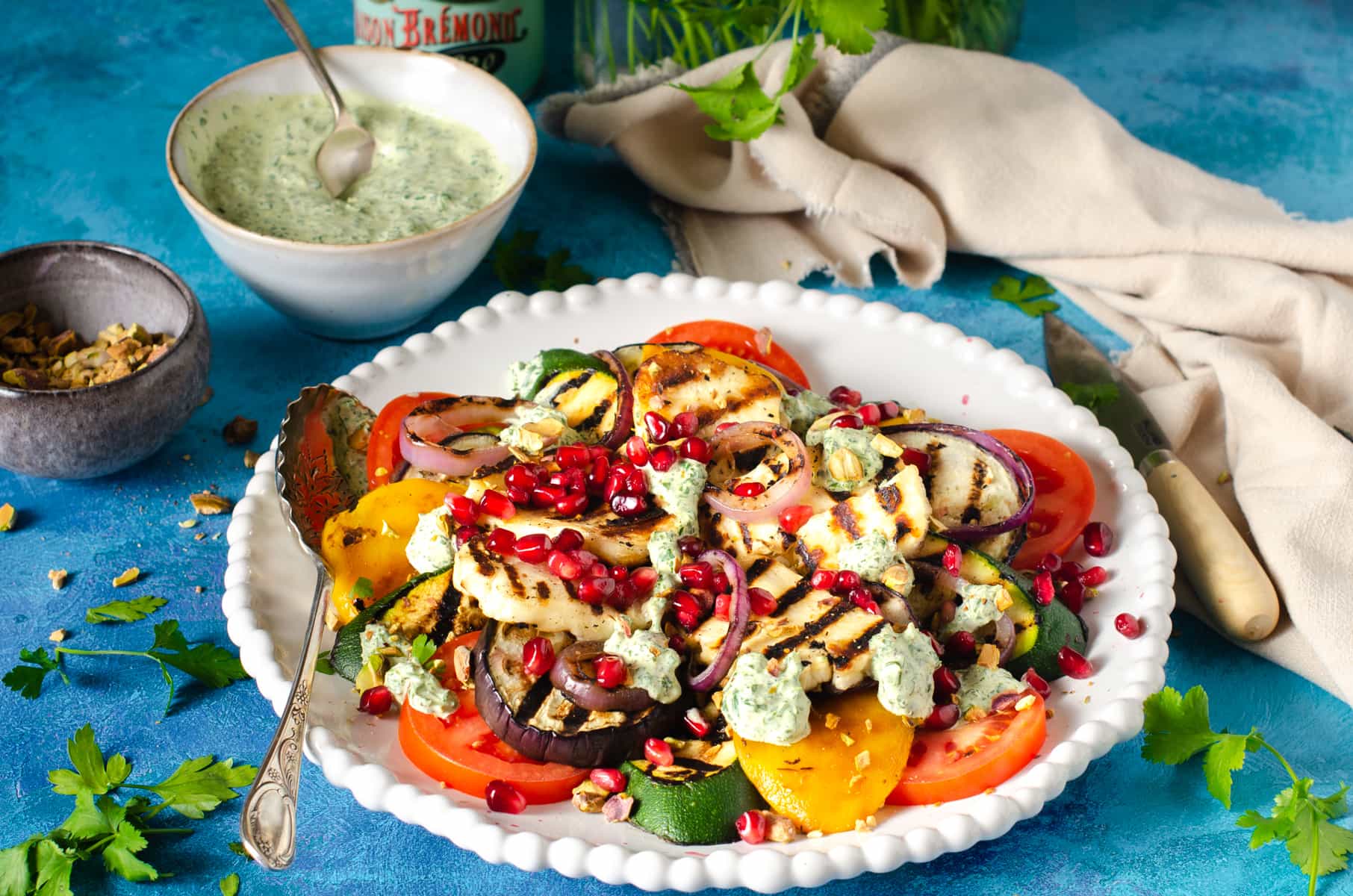 A fresh and vibrant halloumi grilled vegetable salad with a yogurt dressing to the back and fresh toasted pistachios.