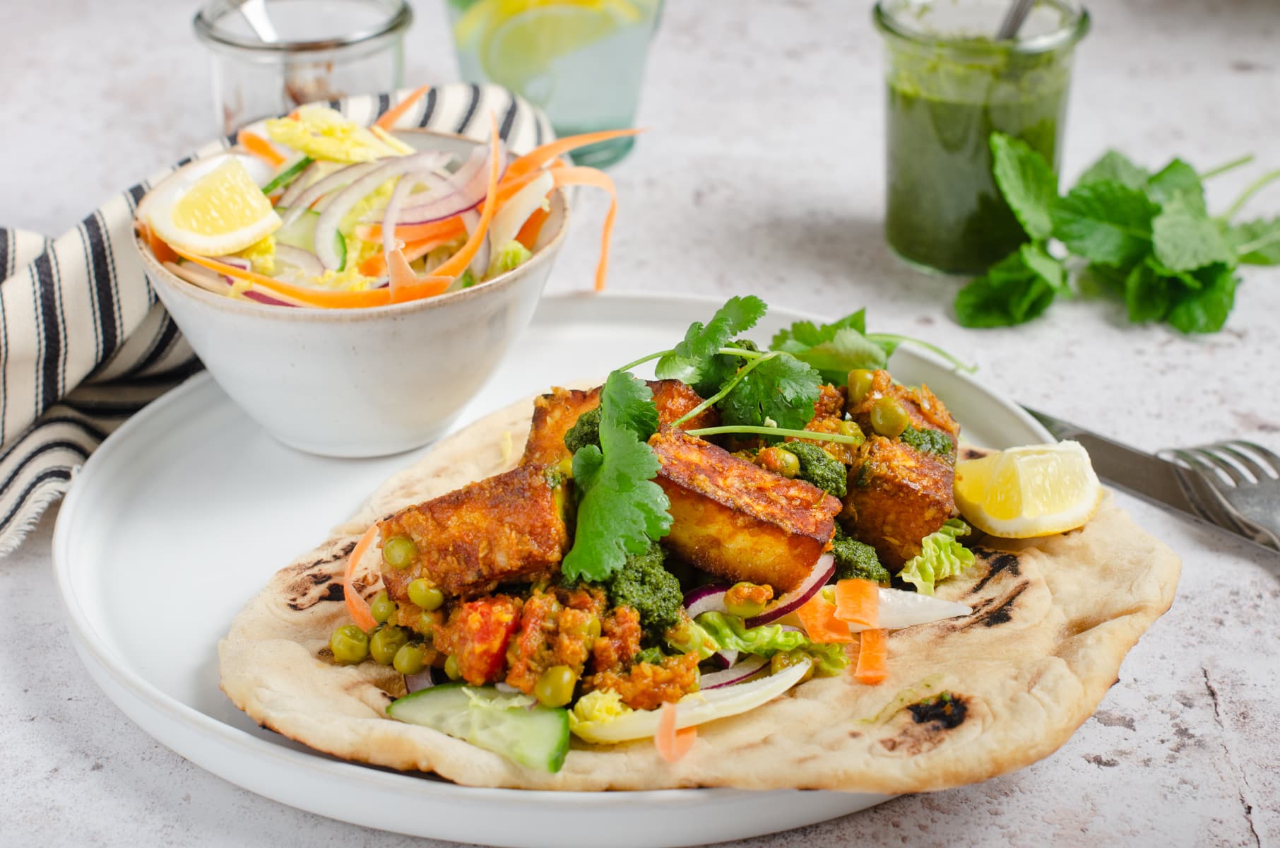 A paneer cheese curry on a top of a flatbread served with a side crunchy salad and green and date chutney.