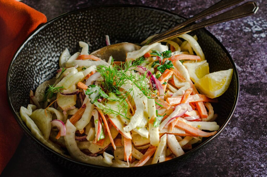 A dark black bowl filled with a fennel and apple coleslaw with an orange linen to the side and servings tongs in the bowl.