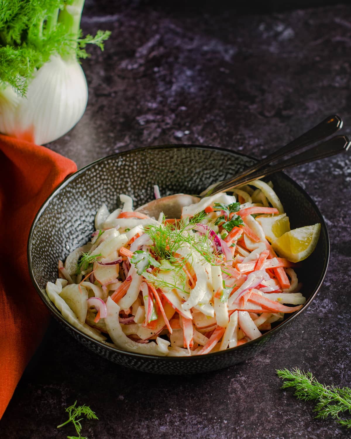 A dark table setting with a bowl of coleslaw filled with fennel, apple and carrot, with sprigs of fennel scattered and an orange coloured linen.
