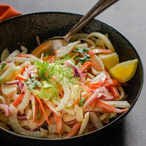 A fresh homemade bowl of fennel slaw garnished with sprigs of fennel and lemon wedges.