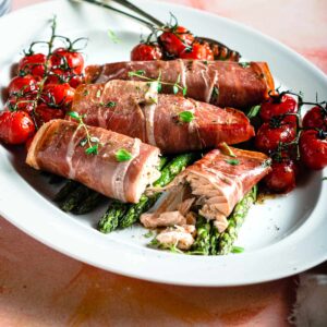 A platter of asparagus, baked salmon in parma ham and oven roasted tomatoes with some glasses to the back ready to be served for dinner.