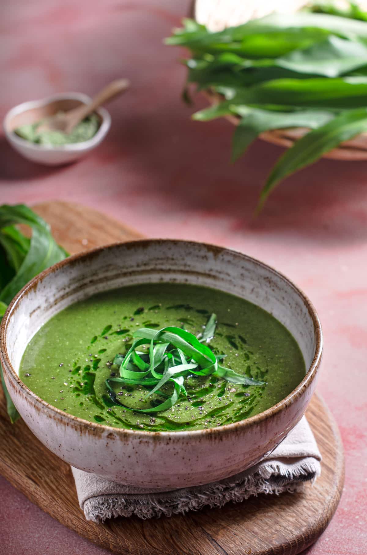 A vibrant green wild garlic soup with fresh picked wild garlic to the back all sat on a pink table top and a wooden board.