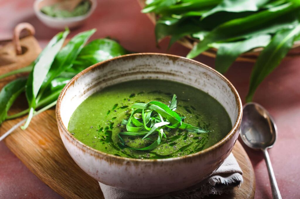A bowl of vibrant green wild garlic soup on a wooden board in a mottled handmade pottery bowl.