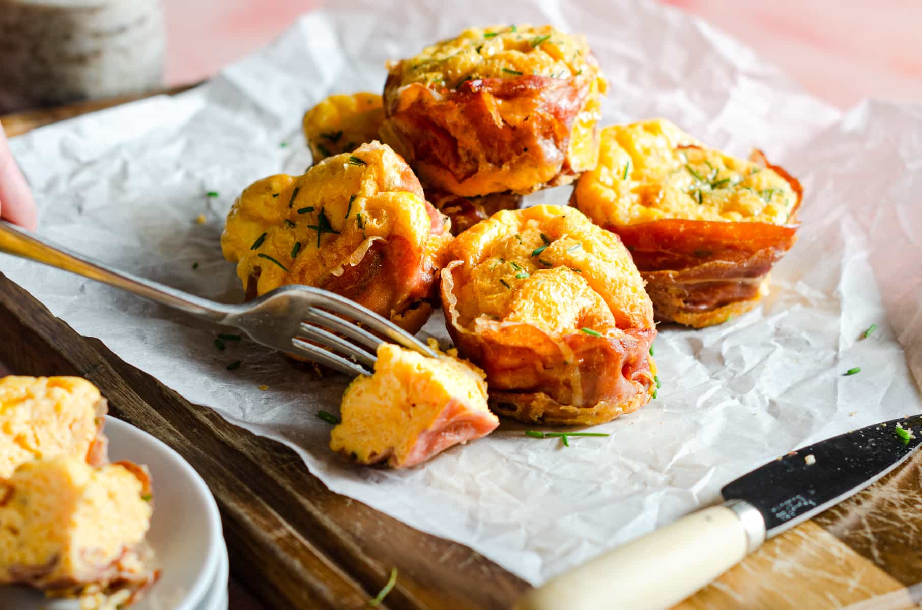 Parma Ham and egg muffins piled on a sheet of baking parchment on a wooden board and a fork cutting into one.