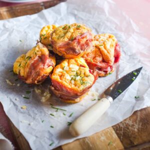 Scrambled egg baked in a muffin tray lined with slices of parma ham and served topped with chopped chives on a wooden board.