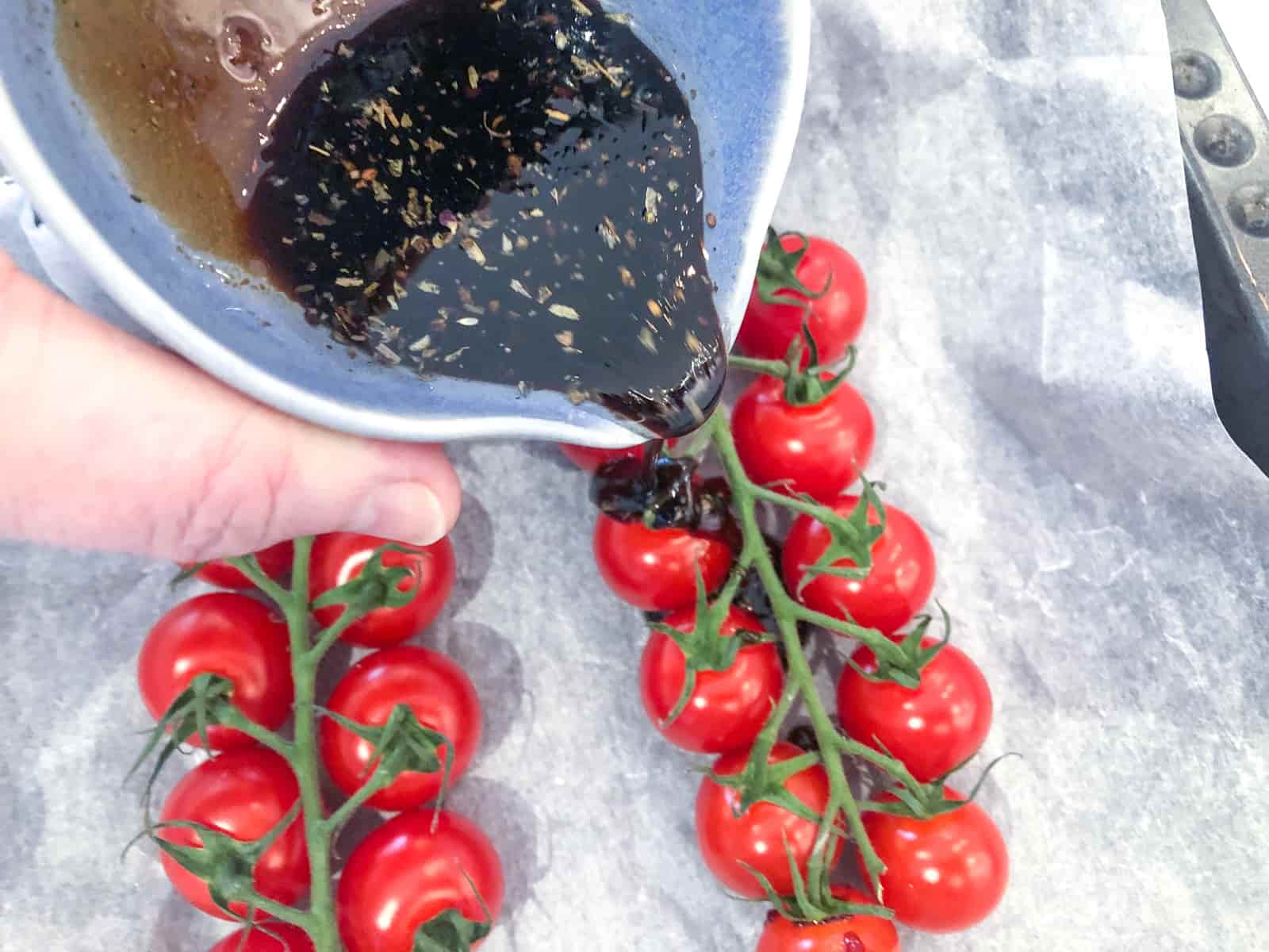 Pouring balsamic dressing over top of vine tomatoes on a baking parchment lined pan.