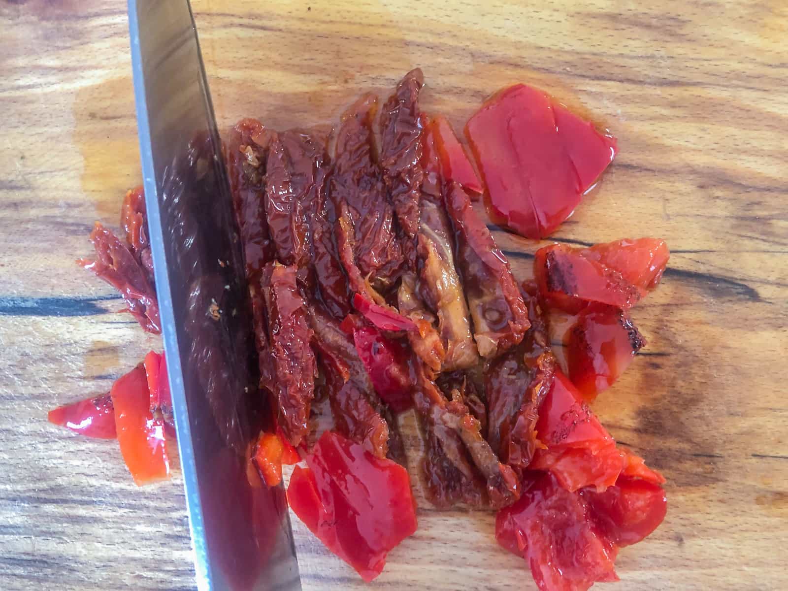 Chopping roasted red peppers and sun-dried tomatoes from a jar into bite sized pieces on a wooden board.