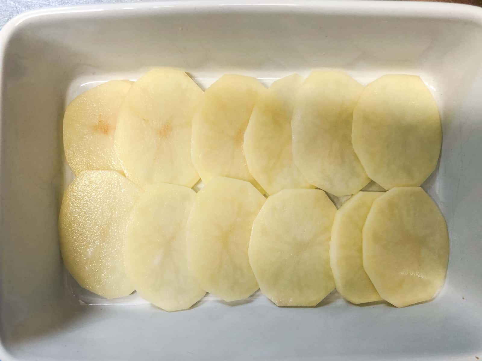 Showing how to prepare a layered boulangere potato dish with slices of potato on the bottom of an oven proof dish.