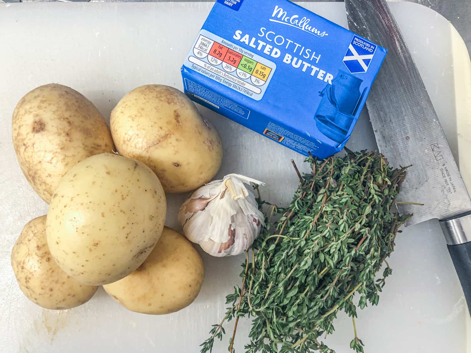 Ingredients to make fondant potatoes on a chopping board consisting of potatoes, fresh thyme, garlic and salted butter.