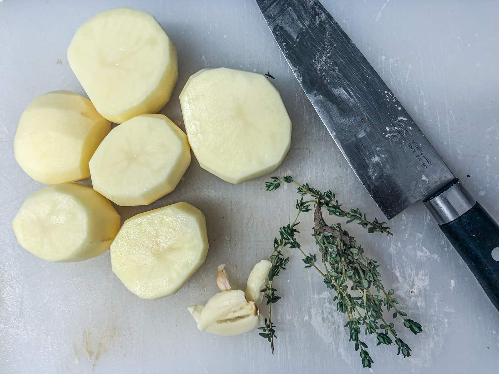 Peeled potatoes, smashed garlic cloves and fresh thyme leaves prepped on a white chopping board with a chefs knife.