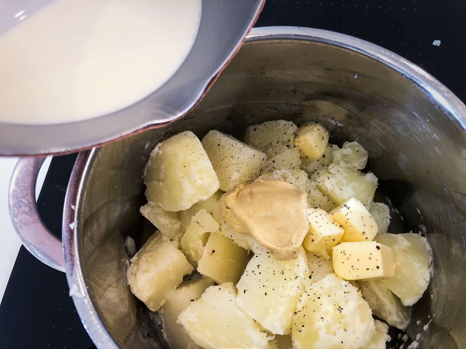 Adding warmed cream to cooked seasoned potatoes with a dollop of mustard before mashing.