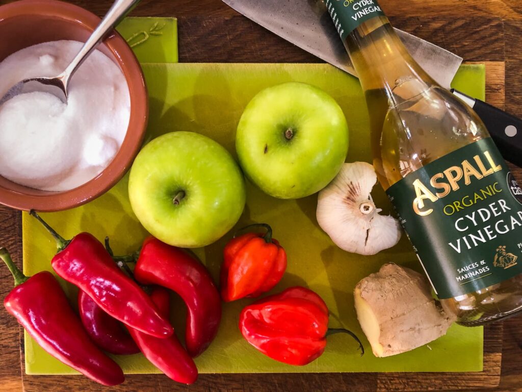 Ingredients for chilli jam on a green chopping board consisting of red chillies, scotch bonnet chillis, cyder vinegar, garlic, ginger, apples and sugar.