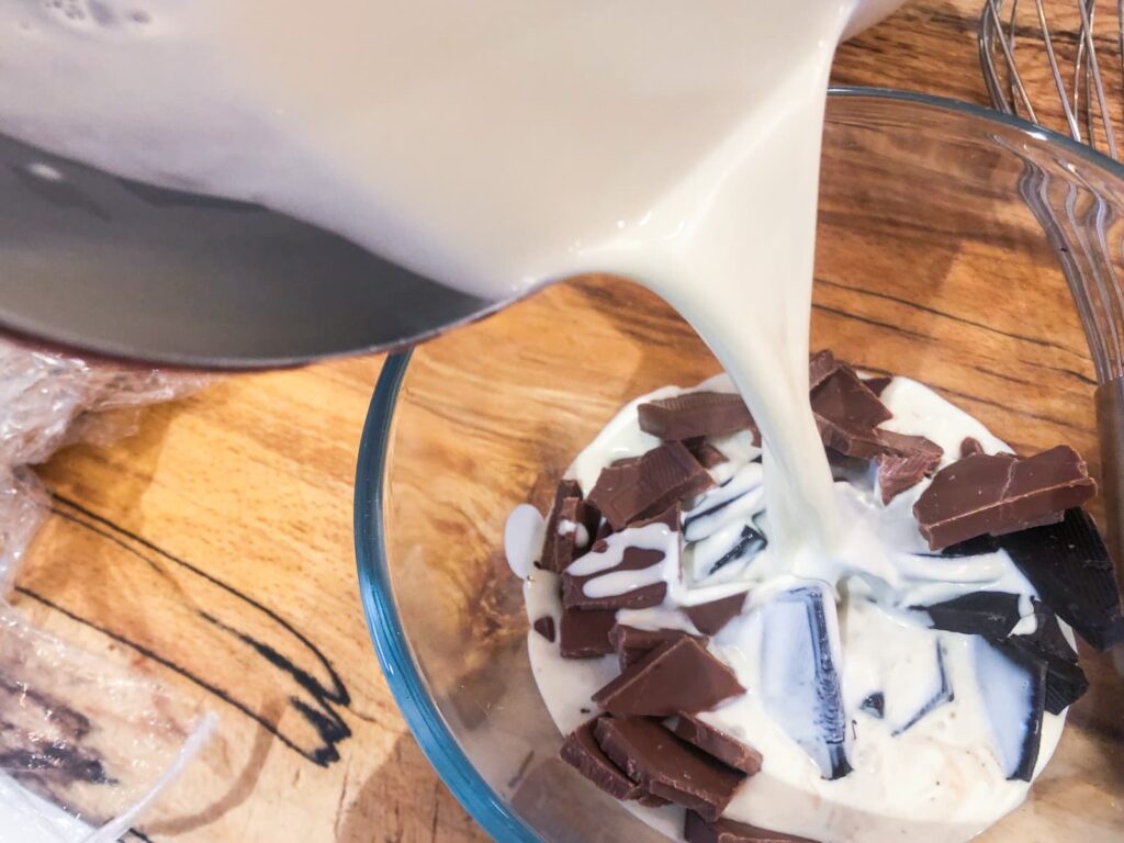 Warmed cream pouring over milk and dark chocolate pieces in a glass bowl to melt the chocolate.