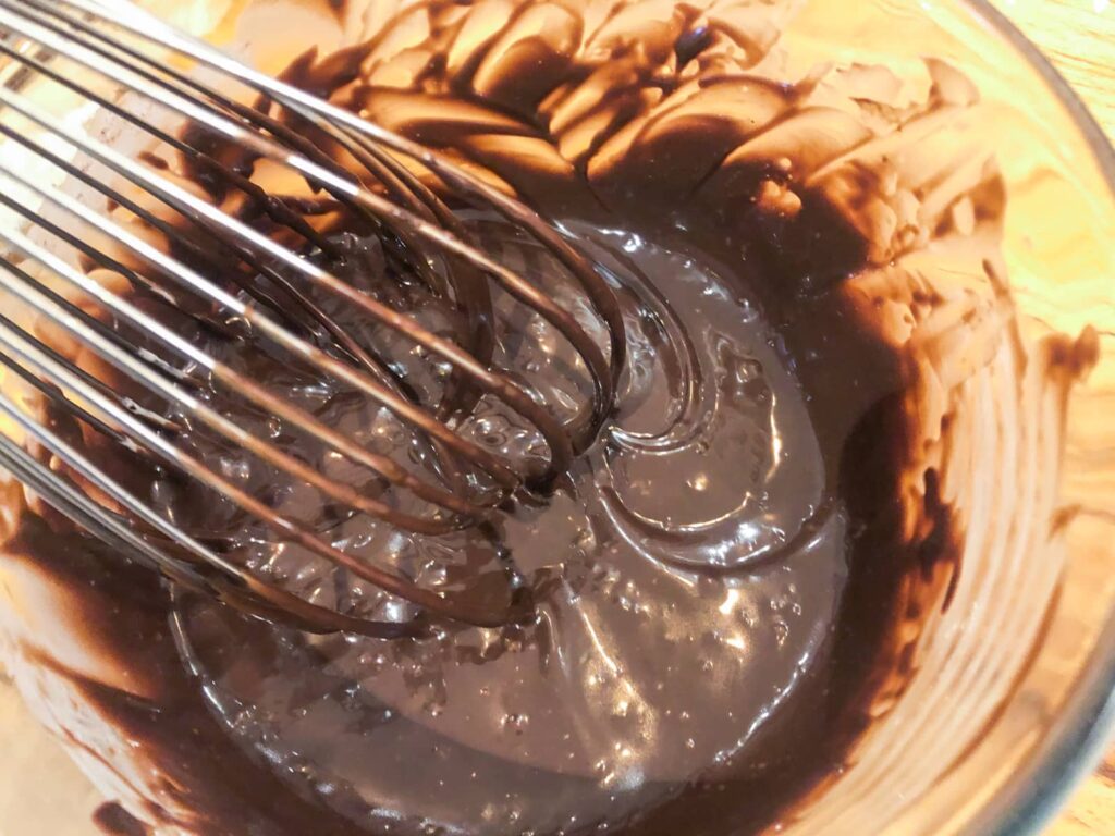 Whisking a smooth chocolate ganache in a glass bowl with a balloon whisk.
