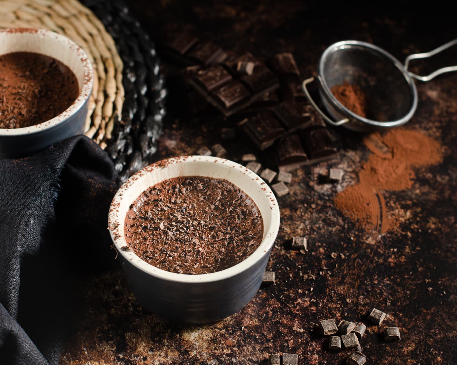 A pot of rich chocolate mousse, topped with chocolate shavings , lots of chocolate pieces scattered around, an old sieve with cocoa powder dusted around on a dark mottled brown marble backdrop.