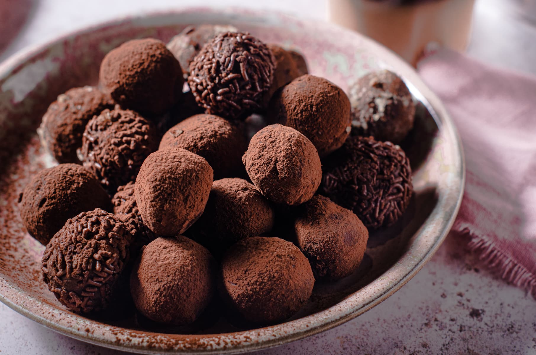 A decadent plate of handmade chocolate truffles dusted in cocoa powder and chocolate sprinkles on a pink mottled plate with a pink linen to the back right.