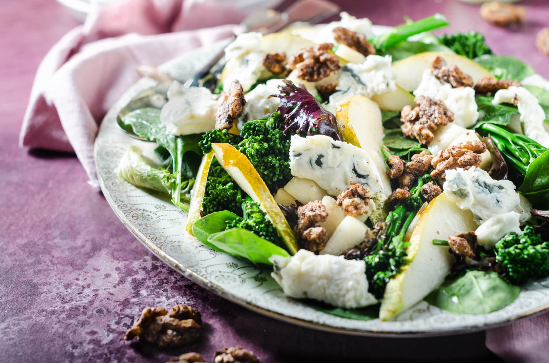 A closeup view of a winter salad consisting of pear, broccoli, spiced walnuts and blue cheese on a gold edged patterned platter sitting on a purple tablecloth.