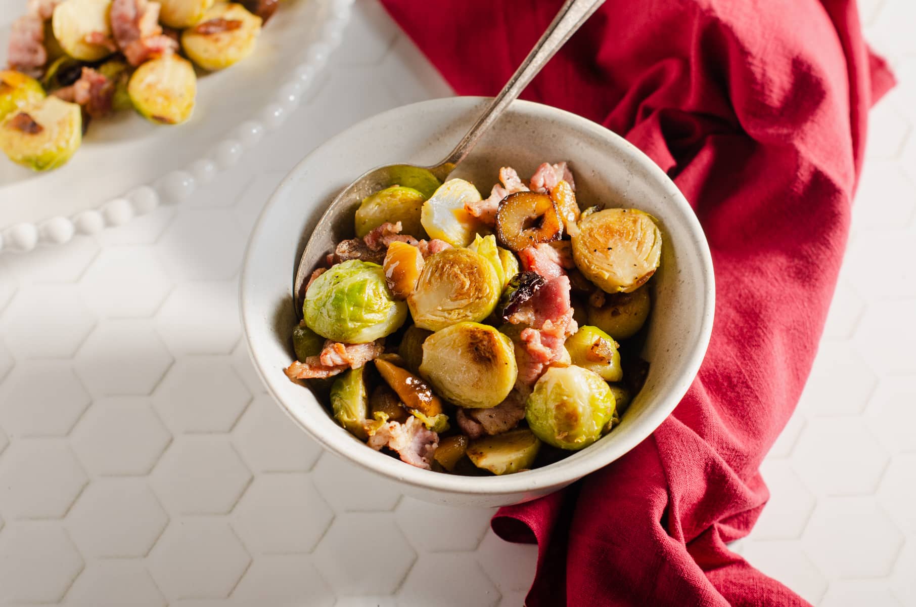A bowl of brussel sprouts cooked with bacon and chestnuts on a white tiled surface and red Christmas linen.