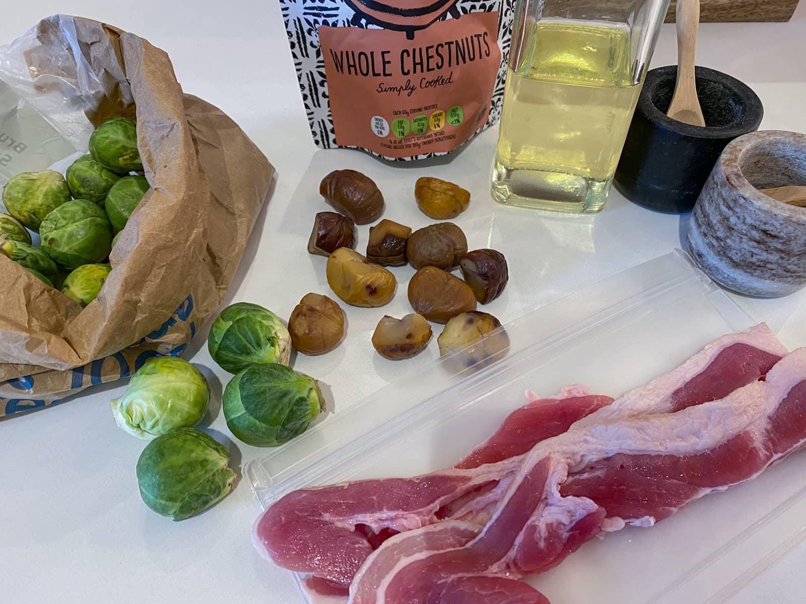 Ingredients on a white surface consisting of sprouts, chestnuts, bacon, oil, salt & pepper.