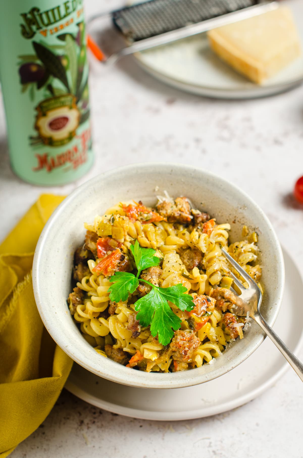 A bowl of creamy pasta with a sausage and tomato sauce on a larger plate with olive oil and parmesan cheese to the back.