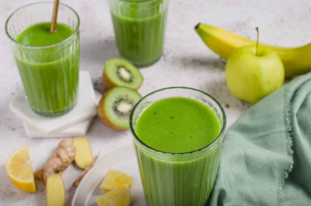 Vibrant green smoothie in glasses with ginger and kiwi to the side.